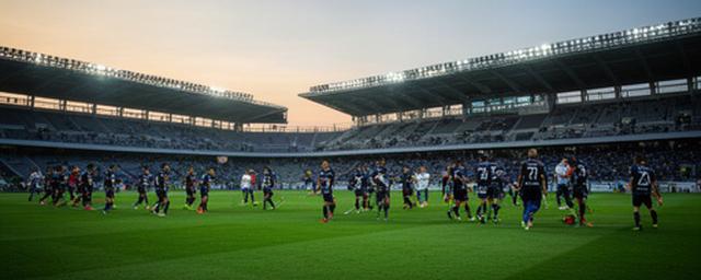 夕方のサッカー専用スタジアムで、緑のピッチを挟んで選手たちが試合に臨む様子